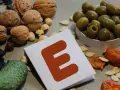 Orzechy, pestki, oliwki i suszone morele wokół kartki z literą "E" symbolizują bogactwo witaminy E.