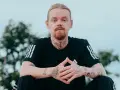 Mężczyzna z rudą brodą i blond włosami, ubrany w czarną koszulkę z trzema paskami. Na szyi i rękach widoczne tatuaże, w tym klucz i napis "kuqe 2115".
