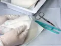 Ręce w rękawiczkach przygotowują jałowe materiały do zmiany opatrunku na ranie czystej. Nożyczki i pęseta leżą obok.