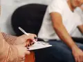 Dłoń pisząca na notatniku, w tle mężczyzna w białej koszulce.