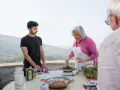 An older woman in an apron cooks greens while a young man and an older man stand nearby on a terrace with a scenic view.