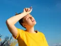 Une jeune femme en t-shirt jaune, le visage marqué par la chaleur, essuie son front sous un ciel bleu intense. La durée d'une insolation est palpable.