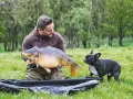 Ein Angler präsentiert stolz einen großen Karpfen, während sein kleiner Hund neugierig zusieht. Ein erfolgreicher Tag beim feedern auf Karpfen.