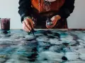 Une artiste crée une **aquarelle sur toile** aux teintes bleues et dorées, ajoutant des touches d'encre avec un compte-gouttes.