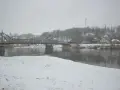 Most na rzece w zimowy dzień, śnieg pada. W tle zabudowania i drzewa. To piękna dx rejestracja krajobrazu.