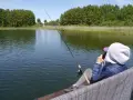 Osoba siedzi na pomoście i łowi ryby na jeziorze Jagodne. W tle las i trzciny.