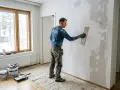 Man spacklar en vägg med spackel och bredspackel. Verktyg och spackelburkar står på golvet.