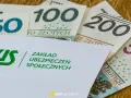 Koperta ZUS z napisem "Zakład Ubezpieczeń Społecznych" na tle polskich banknotów 50, 100, 200 i 500 zł. Symbolizuje świadczenie kompensacyjne.