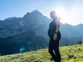 Frau in der Schwangerschaft genießt das Wandern in den Bergen. Die Sonne scheint hinter ihr, während sie auf eine majestätische Bergkulisse blickt.