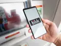 Dłoń trzyma smartfon z logo BLIK przed bankomatem.