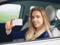 Uśmiechnięta młoda kobieta w samochodzie pokazuje prawo jazdy. Zastanawiasz się, ile kosztuje zdjęcie do prawa jazdy?