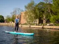 Frau genießt Stand Up Paddling auf einem Fluss, während Menschen am Ufer entspannen.