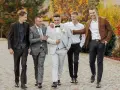 Pięciu mężczyzn w eleganckich garniturach i marynarkach, gotowe stylizacje na wesele, spaceruje po brukowanej ścieżce wśród jesiennej zieleni.