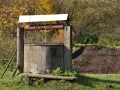Stara studnia kręgowa z drewnianą konstrukcją i metalowym kołowrotem, otoczona jesienną zielenią i stertą ziemi.