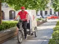 Uśmiechnięty kurier rowerowy DPD w czerwonej koszulce jedzie ulicą z zielonymi drzewami.