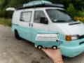 Mała figurka t4 camper w ręce, na tle prawdziwego, turkusowo-białego kampera.