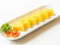 Sześć kawałków puszystego omletu tamago ułożonych na białym talerzu, z gałązką pietruszki i pomarańczowymi dodatkami.