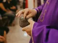 A priest in a purple robe takes ashes from a small bowl, preparing for the first day of Lent prayer.