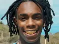 YNW Melly, uśmiechnięty z złotymi zębami, na tle nieba i wzgórz.