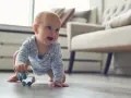 Ein Baby in blauem Sternen-Body krabbelt fröhlich über den Holzboden und greift nach einer bunten Rassel. So erkunden Babys die Welt!
