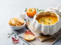 Zupa krem z dyni w naczyniu w kształcie dyni, podana z grzankami i śmietanką. Obok mała dynia i jesienne liście.