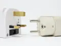 Adaptadores para diferentes tipos de enchufes: uno blanco con tres clavijas doradas y otro beige con dos clavijas plateadas.