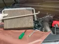 Gloved hand holds car AC evaporator. Tools and engine parts visible. Learn how to make AC colder in car by fixing this component.
