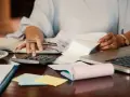 Persona revisa documentos y usa calculadora para el modelo 184. Hay café, portátil y notas adhesivas.
