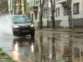 Ciemny samochód wpada w kałużę, powodując potężny **aquaplaning**. Woda rozpryskuje się na boki, a odbicia budynków i drzew tańczą na mokrej nawierzchni.