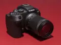 Appareil photo noir Canon EOS R7 avec objectif 18-150 mm, posé sur un fond rouge vif.