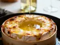 Un mont d'or au four, doré et fondant, est arrosé d'un filet d'huile dans sa boîte en bois.