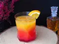 Kolorowy drink z koniakiem, ozdobiony plasterkiem pomarańczy, na tle karafki z bursztynowym alkoholem i kwitnących wrzosów.