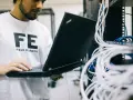 Inżynier w koszulce "FE FIELD ENGINEER" używa laptopa do sprawdzenia **trace route cmd** w serwerowni z plątaniną kabli.