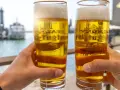 Zwei Hände halten Biergläser mit schaumigem Bier. Im Hintergrund ein Hafen. Ist Bier gesund? Ein Genuss am Wasser.