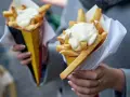 Dwie porcje frytek z majonezem w papierowych opakowaniach, trzymane w dłoniach.
