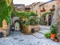 Un vicolo acciottolato con case in pietra ricoperte di edera e fiori. Un uomo cammina verso un arco che si apre su un paesaggio verde. Uno dei tanti posti belli nelle Marche.