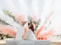 Un couple s'embrasse sous une arche fleurie, entouré de fumée rose et blanche. Ces photos mariage capturent un moment magique.