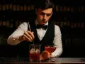 Elegancki barman w kamizelce i muszce miesza drinka z lodem w szklanicy. Obok stoi gotowy koktajl.