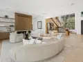 Soggiorno luminoso con camino moderno, divano curvo e poltrone bianche. Scale a vista e grandi finestre definiscono l'arredamento minimal chic.