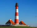 Wysoka, czerwono-biała latarnia morska w Jastarni strzeże wybrzeża. Obok stoją dwa domki z pomarańczowymi dachami.