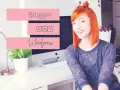 Uśmiechnięta rudowłosa kobieta siedzi przy biurku z komputerem, na którym widnieją napisy "Blogger" i "Wordpress".