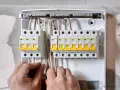 Ręce elektryka podłączają przewody do skrzynki z bezpiecznikami. Widać wlz, czyli rozdzielnicę z wyłącznikami nadprądowymi.