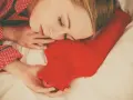 Une femme dort paisiblement, blottie contre une bouillotte rouge. Une astuce pour savoir comment faire une bouillotte avec une serviette pour se réchauffer.