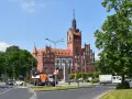 Imponujący ratusz Słupsk z czerwoną cegłą i wieżą góruje nad placem. Wokół drzewa i ruch uliczny.