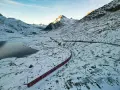 Czerwony pociąg panoramiczny wije się przez ośnieżony górski krajobraz. Słońce oświetla szczyty, a obok jezioro odbija błękitne niebo.