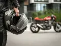 Motociclista in pelle tiene un casco, pronto per misurare la circonferenza della testa per un acquisto perfetto. Moto rossa sfocata sullo sfondo.
