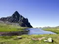Majestuoso pico rocoso se alza sobre un lago sereno y prados verdes en el valle de Tena, con vacas pastando tranquilamente.