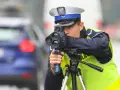 Policjant w kamizelce odblaskowej mierzy prędkość samochodu za pomocą radaru. Trwa dozór policyjny na drodze.
