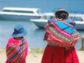 Zwei cholitas escaladoras, gekleidet in bunte Ponchos und Hüte, blicken auf Boote auf einem See.