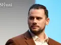 Grzegorz Collins, znany z programów telewizyjnych, w eleganckim brązowym garniturze. Jego wiek jest trudny do określenia, ale prezentuje się dojrzale.